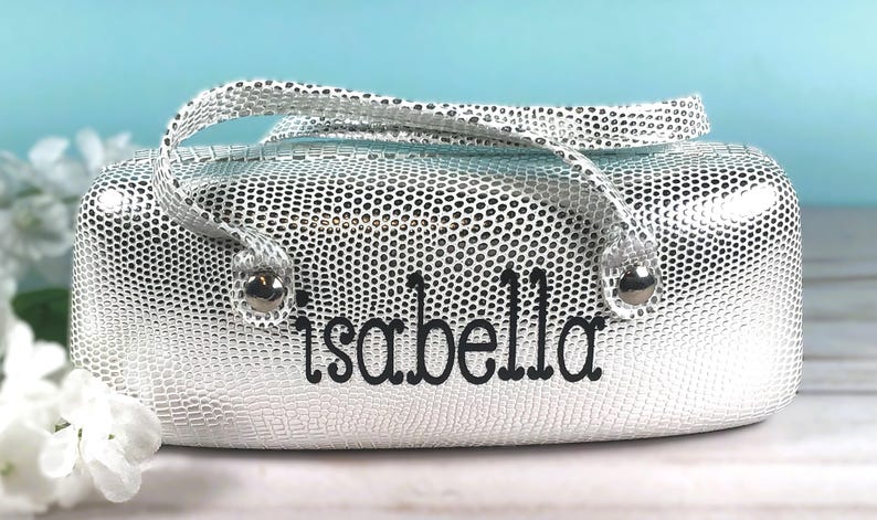 May include: Silver cosmetic bag with a textured, dotted pattern and a matching handle. The name "Isabella" is embroidered in black script on the front. The bag has silver hardware.