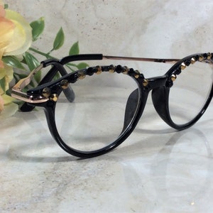 May include: Black-framed spectacles with clear lenses. The upper rims are decorated with small, gold-coloured beads. The temples are rose gold in colour.