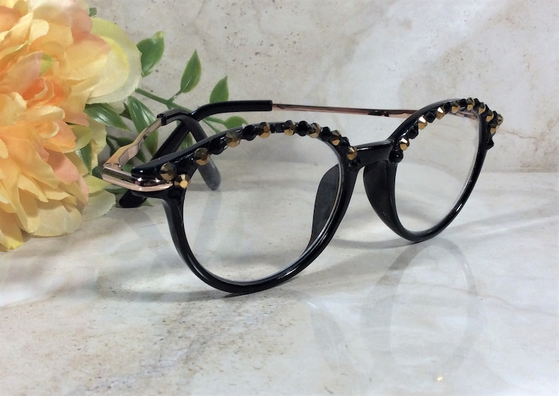 May include: Black-framed spectacles with clear lenses. The upper rims are decorated with small, gold-coloured beads. The temples are rose gold in colour.
