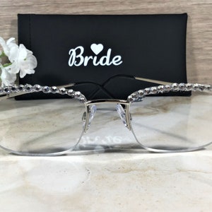 May include: Silver-rimmed eyeglasses with clear lenses, adorned with sparkling rhinestones along the top frame. A black case with the word "Bride" in white script and a heart symbol is behind the glasses. The glasses are a bridal accessory.