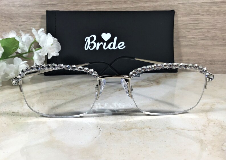 May include: Silver-rimmed eyeglasses with clear lenses, adorned with sparkling rhinestones along the top frame. A black case with the word "Bride" in white script and a heart symbol is behind the glasses. The glasses are a bridal accessory.