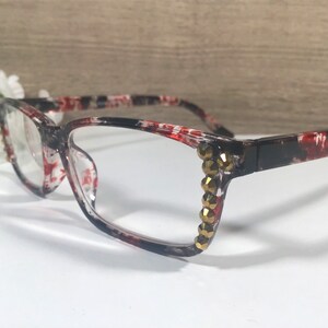 May include: Clear-framed eyeglasses with a red and black floral pattern. The frames are accented with gold-colored rhinestones on the outer corners. The glasses are set against a white surface with a blurred background.