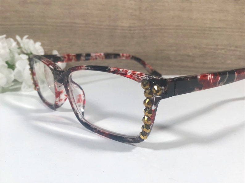 May include: Clear-framed eyeglasses with a red and black floral pattern. The frames are accented with gold-colored rhinestones on the outer corners. The glasses are set against a white surface with a blurred background.