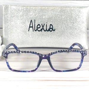 May include: Rectangular eyeglasses with a blue and purple marbled frame, featuring a row of small dark blue rhinestones along the top rim. Behind the glasses sits a silver glittery rectangular case with the name Alexia embroidered in black.