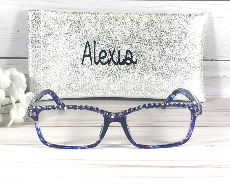 Op de afbeelding: Rechthoekige bril met een blauw en paars gemarmerd montuur, voorzien van een rij kleine donkerblauwe strass-steentjes langs de bovenrand. Achter de bril staat een zilverkleurig glitteretui met de naam Alexia in het zwart geborduurd.