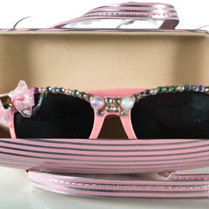 May include: Pink sunglasses with dark lenses, decorated with pearls and rhinestones, are presented in a matching pink striped case. A pink bow is attached to the left side of the sunglasses. The case has a handle and silver trim.