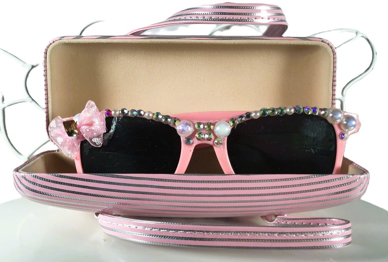 May include: Pink sunglasses with dark lenses, decorated with pearls and rhinestones, are presented in a matching pink striped case. A pink bow is attached to the left side of the sunglasses. The case has a handle and silver trim.