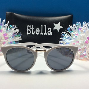 May include: White sunglasses with a gold accent on the bridge. The sunglasses are in a black case with the name "Stella" and a star printed on it.