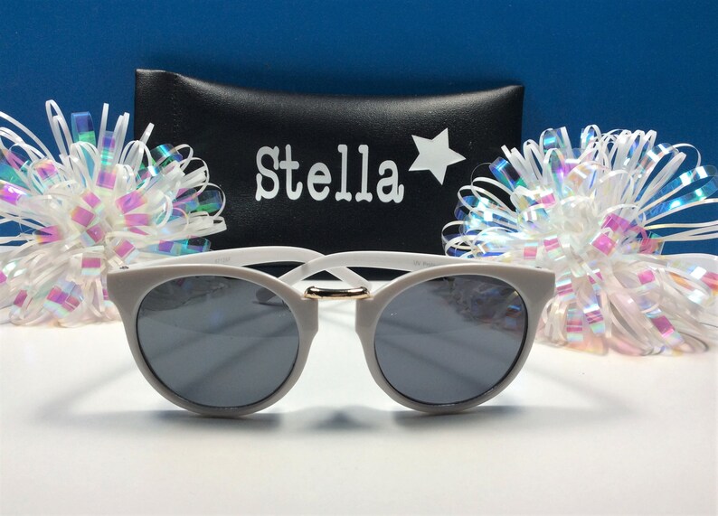 May include: White sunglasses with a gold accent on the bridge. The sunglasses are in a black case with the name "Stella" and a star printed on it.
