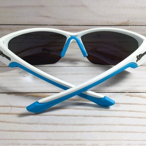 May include: Sport sunglasses featuring a white frame with bright blue accents on the temple tips and nose pads. The lenses are dark grey and wrap around the face. The word Xtreme is printed in black on both sides of the white frame temples.