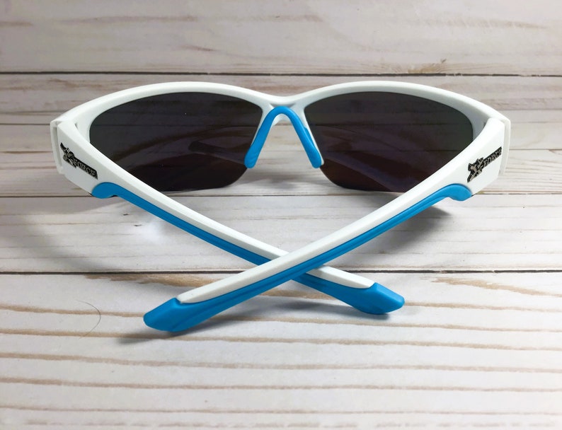 May include: Sport sunglasses featuring a white frame with bright blue accents on the temple tips and nose pads. The lenses are dark grey and wrap around the face. The word Xtreme is printed in black on both sides of the white frame temples.