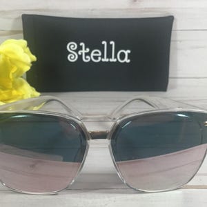 May include: Clear framed sunglasses with rose gold mirrored lenses. The sunglasses are sitting on a white surface with a black case behind them. The case has the word "Stella" printed on it in white.