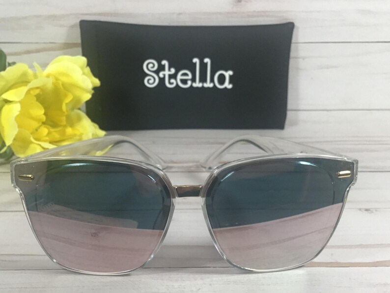 May include: Clear framed sunglasses with rose gold mirrored lenses. The sunglasses are sitting on a white surface with a black case behind them. The case has the word "Stella" printed on it in white.
