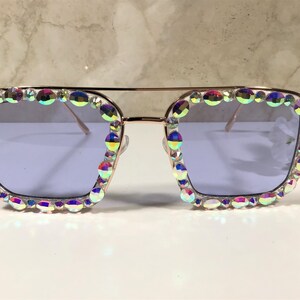 Peut inclure: Une paire de lunettes de soleil carr&eacute;es avec une monture dor&eacute;e et des strass iris&eacute;s autour des verres. Les verres sont de couleur violet clair.