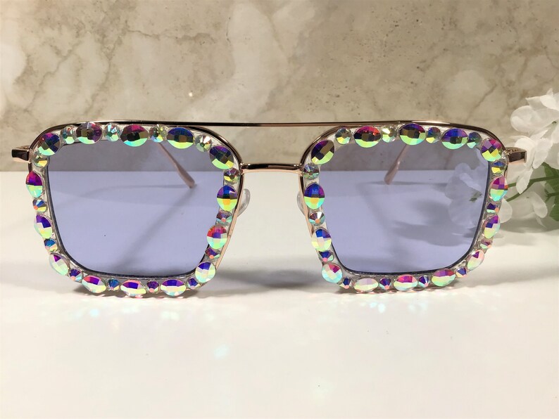 Peut inclure: Une paire de lunettes de soleil carr&eacute;es avec une monture dor&eacute;e et des strass iris&eacute;s autour des verres. Les verres sont de couleur violet clair.