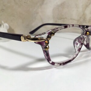 Puede incluir: Gafas transparentes con un patr&oacute;n moteado morado y blanco en las monturas. Las patillas son de color morado oscuro y las bisagras doradas. Adornos dorados en la montura.