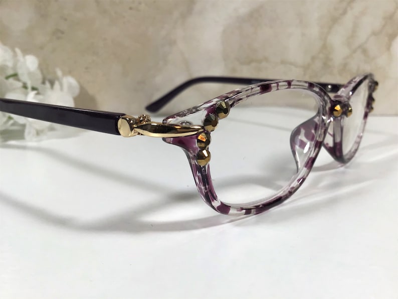 Puede incluir: Gafas transparentes con un patr&oacute;n moteado morado y blanco en las monturas. Las patillas son de color morado oscuro y las bisagras doradas. Adornos dorados en la montura.