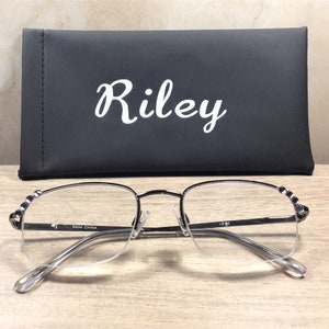 May include: Silver-framed reading glasses with clear lenses and a black case. The case has the name "Riley" in white script. The glasses have a half-rim design and decorative accents on the temples. The glasses are resting on a wooden surface.