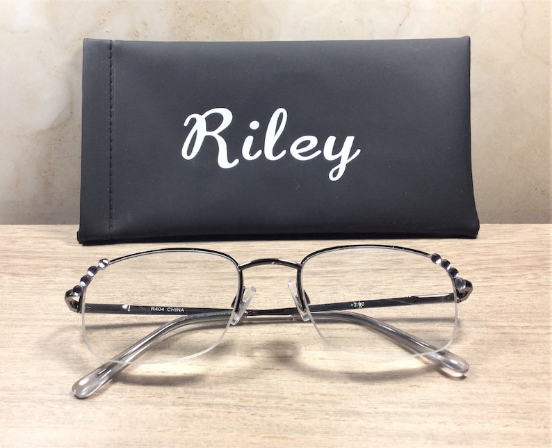 May include: Silver-framed reading glasses with clear lenses and a black case. The case has the name "Riley" in white script. The glasses have a half-rim design and decorative accents on the temples. The glasses are resting on a wooden surface.