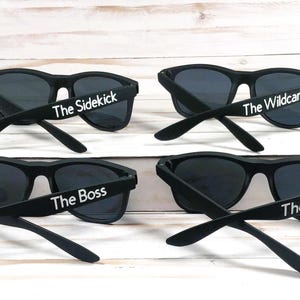 Op de afbeelding: Vier paar zwarte zonnebrillen met donkere glazen. Elk paar is voorzien van witte tekst op de brilpoot. De tekst luidt The Sidekick, The Wildcard, The Boss en The Bud, respectievelijk op de vier afzonderlijke brillen in de afbeelding.