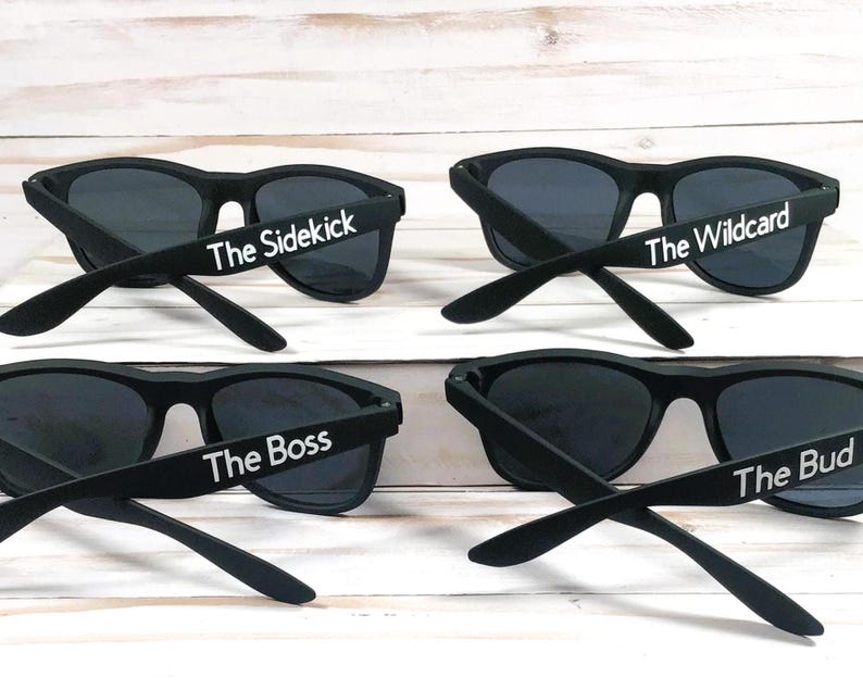 Op de afbeelding: Vier paar zwarte zonnebrillen met donkere glazen. Elk paar is voorzien van witte tekst op de brilpoot. De tekst luidt The Sidekick, The Wildcard, The Boss en The Bud, respectievelijk op de vier afzonderlijke brillen in de afbeelding.