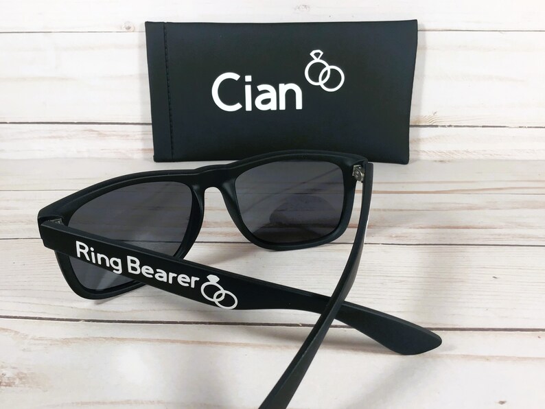 May include: Black sunglasses with the text "Ring Bearer" and two interlocking rings on the arm. The sunglasses come with a black case that has the name "Cian" and two interlocking rings printed on it.