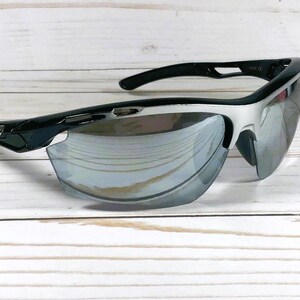 May include: Sport sunglasses featuring a black and silver frame with a wrap-around design. The lenses are reflective and mirrored, showing a silver tint. The temples are black with a small logo detail on the side, and the frame has a sleek, aerodynamic shape.