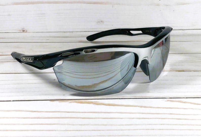 May include: Sport sunglasses featuring a black and silver frame with a wrap-around design. The lenses are reflective and mirrored, showing a silver tint. The temples are black with a small logo detail on the side, and the frame has a sleek, aerodynamic shape.