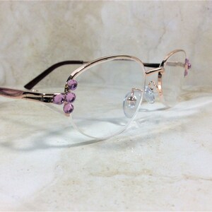 Pu&ograve; includere: Occhiali color oro rosa con lenti trasparenti. Le montature presentano gruppi decorativi di gemme viola chiaro sulle aste e sul lato destro. Le aste sono di colore marrone scuro.