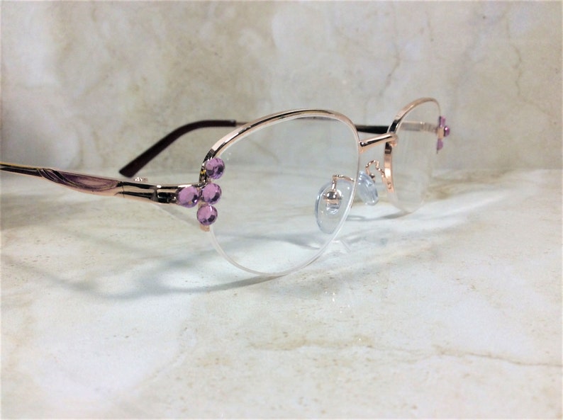 Pu&ograve; includere: Occhiali color oro rosa con lenti trasparenti. Le montature presentano gruppi decorativi di gemme viola chiaro sulle aste e sul lato destro. Le aste sono di colore marrone scuro.