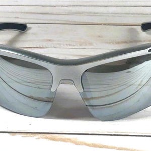 May include: Sport sunglasses featuring a silver frame and reflective mirrored lenses. The design includes a wrap-around style with dark grey rubberized temple tips and nose pads, providing a sleek look with ventilation ports along the upper frame.