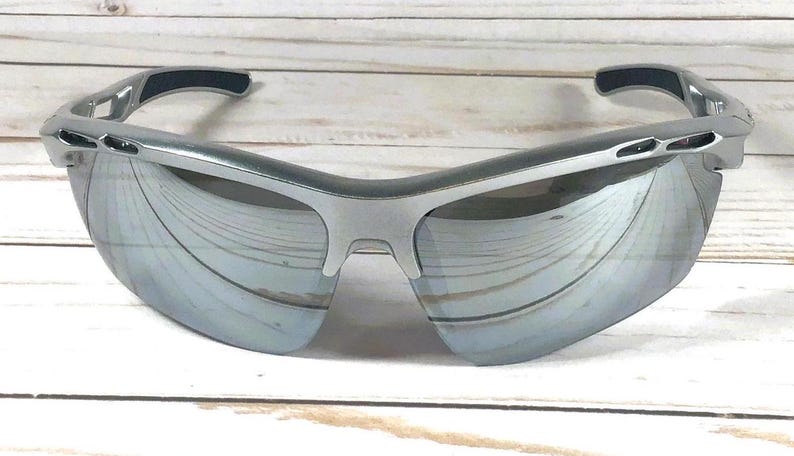 May include: Sport sunglasses featuring a silver frame and reflective mirrored lenses. The design includes a wrap-around style with dark grey rubberized temple tips and nose pads, providing a sleek look with ventilation ports along the upper frame.