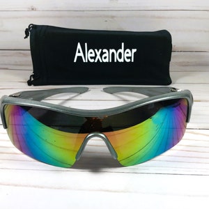 May include: Silver-framed sunglasses with iridescent rainbow lenses. The sunglasses are on a white wooden surface. The sunglasses come with a black carrying case that says "Alexander" on it.