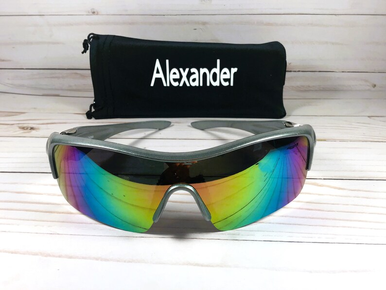 May include: Silver-framed sunglasses with iridescent rainbow lenses. The sunglasses are on a white wooden surface. The sunglasses come with a black carrying case that says "Alexander" on it.