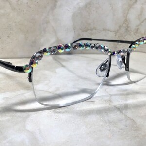 May include: Black and silver-rimmed eyeglasses with clear lenses. The top of the frames is adorned with iridescent, sparkling rhinestones. The glasses have a half-rim design and nose pads.