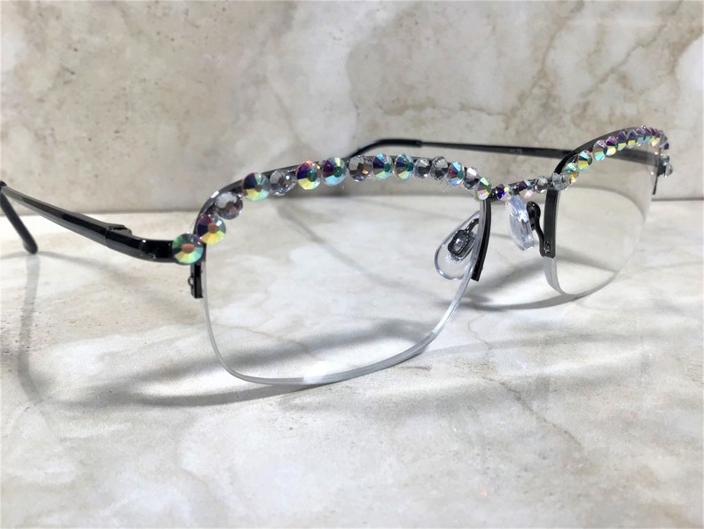 May include: Black and silver-rimmed eyeglasses with clear lenses. The top of the frames is adorned with iridescent, sparkling rhinestones. The glasses have a half-rim design and nose pads.