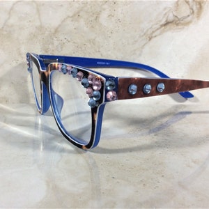 May include: Blue and tortoiseshell reading glasses with clear lenses. The frames are embellished with pink and blue rhinestones. The arms have a brown marbled pattern and blue accents. The glasses are set against a light-colored background.