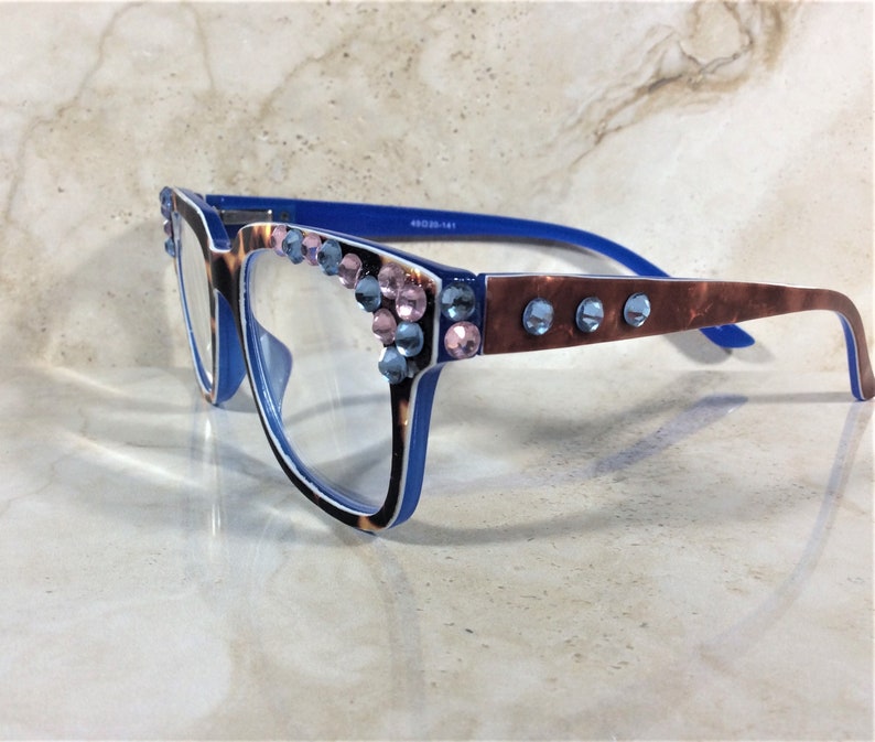 May include: Blue and tortoiseshell reading glasses with clear lenses. The frames are embellished with pink and blue rhinestones. The arms have a brown marbled pattern and blue accents. The glasses are set against a light-colored background.