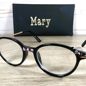 May include: Black cat-eye glasses with pink gemstone embellishments and gold-toned arms. A black case with the name "Mary" in gold lettering is visible behind the glasses, resting on a wooden surface.