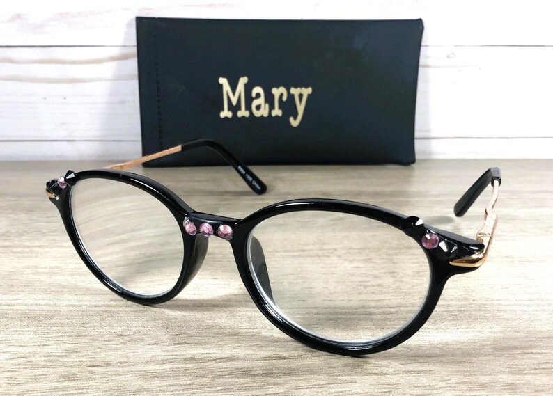 May include: Black cat-eye glasses with pink gemstone embellishments and gold-toned arms. A black case with the name "Mary" in gold lettering is visible behind the glasses, resting on a wooden surface.