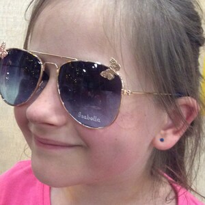 Op de afbeelding: Een jong persoon draagt een gouden aviator zonnebril met vlinderaccenten. De zonnebril heeft donkere glazen en het woord "Isabella" is gegraveerd op de rechterlens.