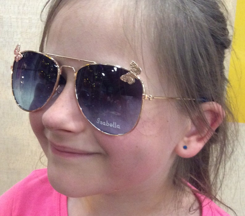 Op de afbeelding: Een jong persoon draagt een gouden aviator zonnebril met vlinderaccenten. De zonnebril heeft donkere glazen en het woord "Isabella" is gegraveerd op de rechterlens.
