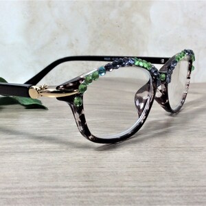May include: Reading glasses with a tortoiseshell pattern frame, adorned with green and blue rhinestones along the top edge. The glasses have black arms and a gold-colored hinge. The lenses are clear.
