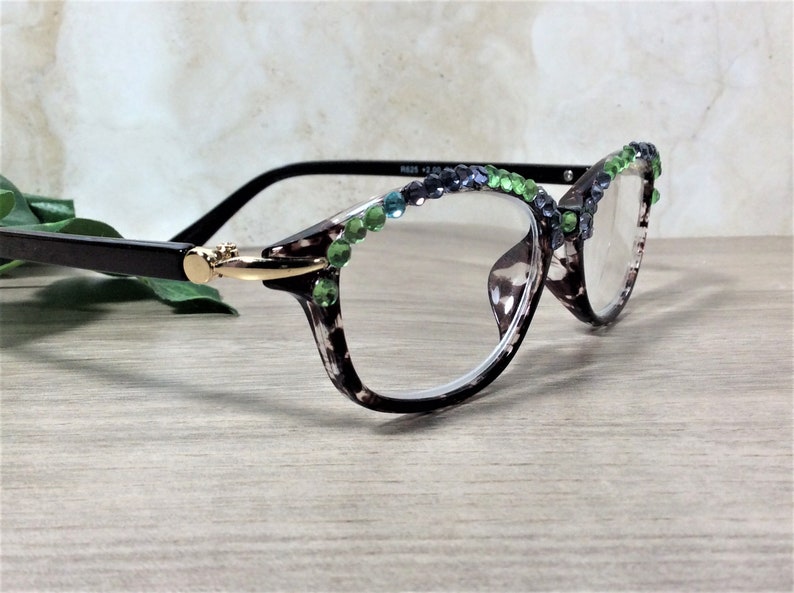 May include: Reading glasses with a tortoiseshell pattern frame, adorned with green and blue rhinestones along the top edge. The glasses have black arms and a gold-colored hinge. The lenses are clear.