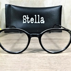 K&ouml;nnte beinhalten: Schwarze Brille mit runden Rahmen, die an den &auml;u&szlig;eren Ecken mit kleinen dunklen Zierkristallen besetzt ist. Hinter der Brille befindet sich ein schwarzes rechteckiges Etui mit dem Wort Stella in wei&szlig;en Buchstaben auf der Vorderseite.