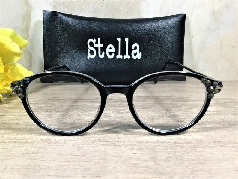 K&ouml;nnte beinhalten: Schwarze Brille mit runden Rahmen, die an den &auml;u&szlig;eren Ecken mit kleinen dunklen Zierkristallen besetzt ist. Hinter der Brille befindet sich ein schwarzes rechteckiges Etui mit dem Wort Stella in wei&szlig;en Buchstaben auf der Vorderseite.