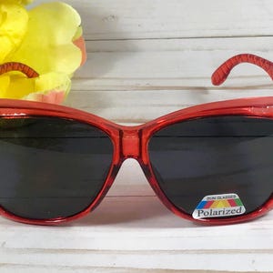 May include: Red sunglasses with a cat-eye shape and dark lenses. The frames are embellished with small rhinestones along the top edge. The sunglasses have a polarized lens sticker.