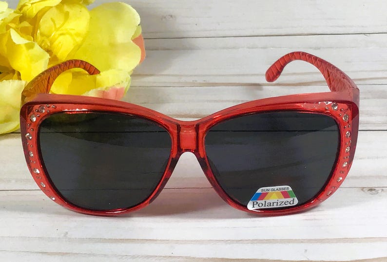 May include: Red sunglasses with a cat-eye shape and dark lenses. The frames are embellished with small rhinestones along the top edge. The sunglasses have a polarized lens sticker.