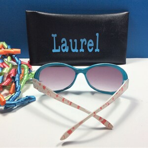 May include: Teal sunglasses with pink tinted lenses and a white frame with a red heart pattern. The sunglasses are folded and the case is black with the name "Laurel" written in blue.