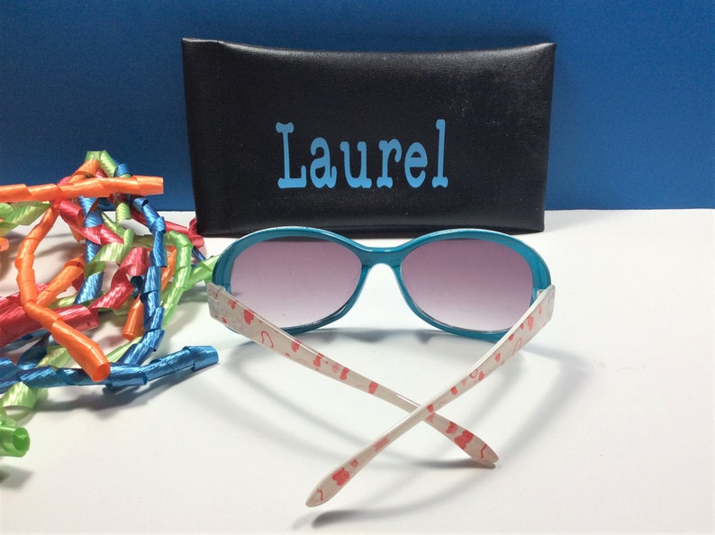 May include: Teal sunglasses with pink tinted lenses and a white frame with a red heart pattern. The sunglasses are folded and the case is black with the name "Laurel" written in blue.
